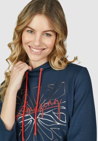 Marine hoodie met rode trekkoorden, voorzien van een bloemig grafisch ontwerp in metallic goud en een vetgedrukte, gestileerde tekst die "Amazing" vermeldt.