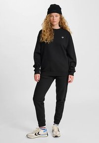 Sudadera negra con puños acanalados, combinada con pantalones deportivos negros. Se lleva con un gorro negro y zapatillas crema con detalles en azul y negro.