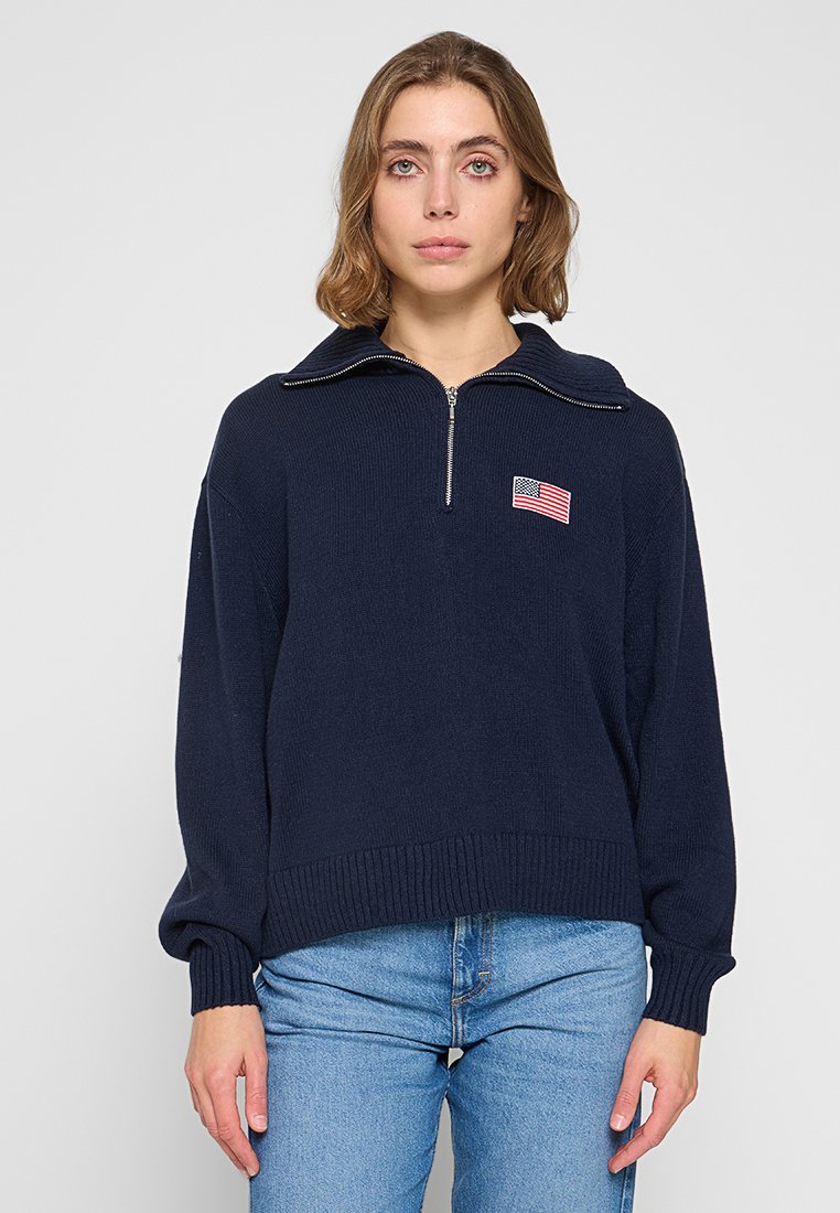 Suéter de punto de color azul marino oscuro, con media cremallera y puños acanalados. Parche bordado de la bandera estadounidense en el pecho.