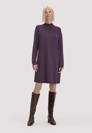 Frau mit glattem weißen Haar, die ein langärmeliges lila Kleid und dunkelbraune kniehohe Stiefel mit Absatz trägt, steht vor einem einfarbigen Hintergrund.