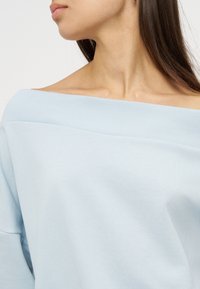 Femme portant un pull bleu clair à épaules dénudées, mettant en valeur le décolleté et les clavicules, avec de longs cheveux bruns partiellement visibles.
