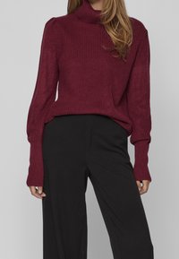 Femme portant un pull en maille bordeaux à manches longues rentré dans un pantalon taille haute noir à jambes larges, sur fond gris uni.