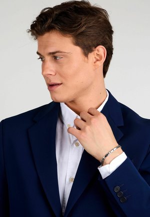 Giacca di abito blu navy con tre bottoni neri, camicia bianca sotto e braccialetto in argento sul polso.