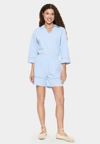 Gaiši zils īsām piedurknēm kombinzons ar V veida kakla izgriezumu un elastīgu jostasvietu, kam ir dekoratīva izšūšana uz piedurknēm un šortiem.