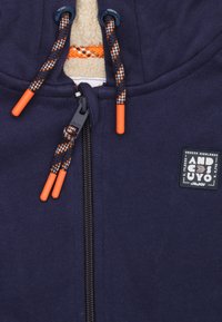 Marineblauwe zip-up hoodie met een textuur fleece interieur, voorzien van oranje ritsstoppers, gevlochten trekkoorden en een logo patch op de borst.