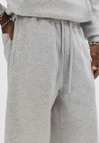 Close-up of person wearing light grey sweatpants with drawstring waist and hands in pockets, showing fabric texture and silver bracelets.