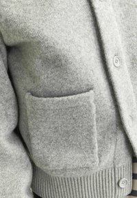 Grå ulltröja med strukturerad yta, framknappar, ribbad nederkant och en stor ficka på vänster sida.