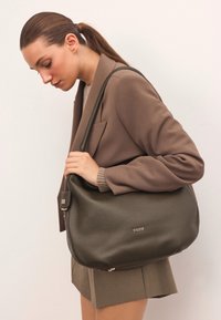 Olíva zöld bőr kézitáska sima felülettel, cipzáras záródással és aranyszínű logóval. A modellen barna blézer és bézs kötött pulóver található.