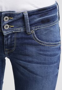Jeans de mezclilla azul con un diseño ajustado, dos botones plateados con marca en la cinturilla y costuras amarillas visibles como acentos.