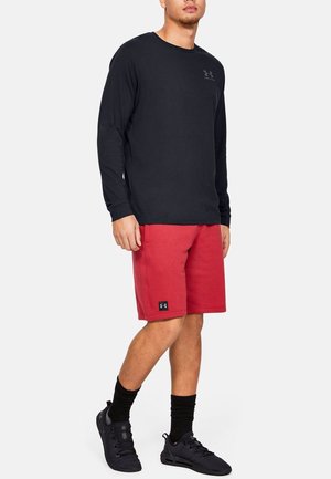 Zwarte lange mouwen shirt, rode sportieve shorts en zwarte schoenen, met een logo op het shirt en een label op de shorts; casual sportieve outfit.