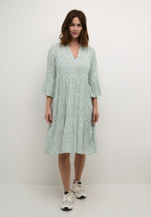 Mintgrünes Blumenkleid mit V-Ausschnitt, dreiviertellangen ausgestellten Ärmeln und gestuftem Rock. Kombiniert mit weißen und schwarzen Sneakers.
