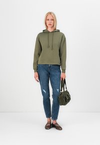 Olíva zöld kapucnis pulóver, húzózsinórral, elhasználódott kék farmernadrággal és fekete csíkos loafers-szel párosítva. Sötétzöld táska a kezében.