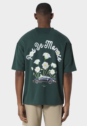 Mann som bærer mørkegrønn skjorte med hvite blomster og svart klassisk bilgrafikk, beige shorts, står med ryggen til på ensfarget bakgrunn.