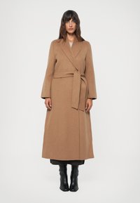 Manteau long en laine beige avec un design enveloppant, ceinture à nouer et manches larges. Porté sur des vêtements sombres et associé à des bottes noires jusqu'aux genoux.