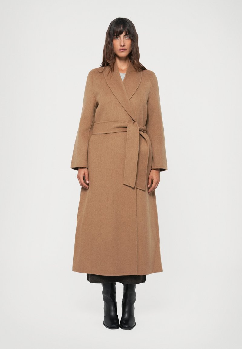 Manteau long en laine beige avec un design enveloppant, ceinture à nouer et manches larges. Porté sur des vêtements sombres et associé à des bottes noires jusqu'aux genoux.