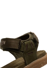 Olivgrön sandal med en mjuk, strukturerad tygöversida, justerbar rem och tan-färgad dämpad sula. Utmärkande sömnads- och varumärkesdetaljer.