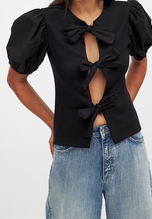 Haut noir avec manches bouffantes, doté de trois fermetures nouées en forme de nœud à l'avant. Confectionné dans un tissu lisse, il présente une coupe ajustée et un détail découpé.