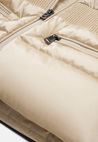 Beige polstret jakke med en glat satinoverflade, vertikale ribbede detaljer og en lynlåslomme med sorte metaldele.