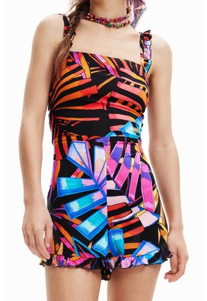 Femme portant une combinaison sans manches avec des motifs géométriques vibrants en bleu, rose, orange et noir, associée à un collier coloré en perles.