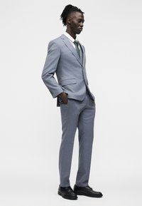 Άνδρας με ανοιχτό γκρι κοστούμι, λευκό πουκάμισο, πράσινη γραβάτα, μαύρα παπούτσια, στέκεται με τα χέρια στις τσέπες σε απλό λευκό φόντο.