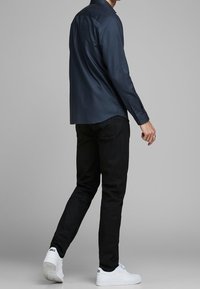 Man som bär en mörkblå långärmad skjorta, svarta byxor och vita ASICS-sneakers som går mot en grå bakgrund.