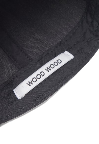 Juodas audinys su etikete "WOOD WOOD" juodu tekstu, įsiūtas į tekstūruoto tamsaus daikto siūlę. Konkretus dizainas, jokių papildomų akcentų.