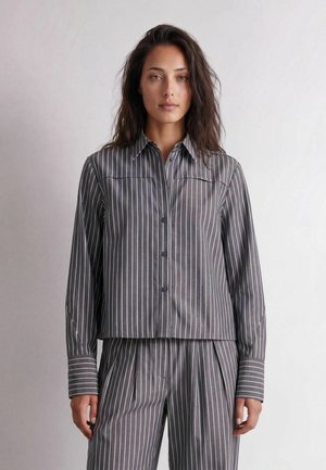 Femme aux longs cheveux foncés portant une chemise grise à fines rayures boutonnée et un pantalon plissé assorti, debout devant un fond clair uni.