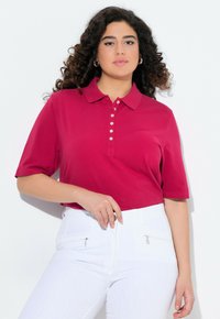 Polo a maniche corte con colletto in magenta vivace, dotato di bottoniera. Indossato con pantaloni bianchi con tasche a zip.