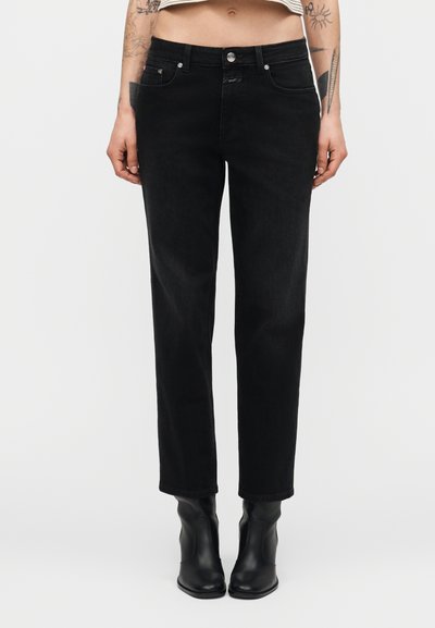 Femme portant un jean noir taille haute à jambes droites et des bottines en cuir noir, avec des tatouages visibles sur les bras et le ventre découvert.