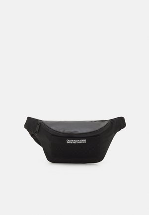 Banane noire avec une section supérieure lisse, une poche avant en mesh et un logo imprimé sur le côté. Fermeture éclair et sangle ajustable.