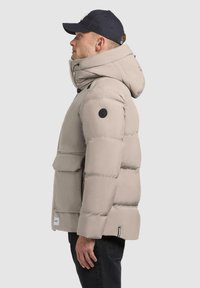 Beige puffer jacket with a large hood and front pocket. Features a logo patch on the arm. Soft, quilted texture with a relaxed fit.
