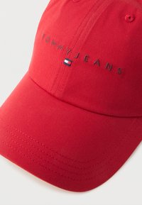 Κόκκινο καπέλο μπέιζμπολ με την επιγραφή "TOMMY JEANS" κεντημένη πάνω από ένα μικρό ορθογώνιο λογότυπο σε κόκκινο, λευκό και μπλε χρώμα στην μπροστινή πάνελ.