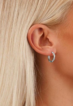 Silberne Creolen mit strukturierter Oberfläche, glänzender Oberfläche und kreisförmiger Form, die am Ohr eines Modells getragen werden.