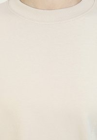Lichtbeige sweatshirt met een geribde kraag, zachte textuur en minimale naden. De stof lijkt glad en lichtgewicht.