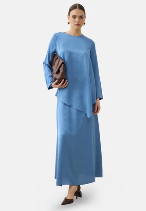 Donna che indossa un lungo vestito di raso blu, tiene una grande pochette marrone e indossa tacchi scuri e orecchini d’oro con pietre rosse.