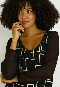 Top negro con diseño de escote en V, que presenta mangas largas transparentes y patrones geométricos dorados metálicos sobre una base sólida.