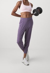 Jasnoróżowy stanik sportowy z białym logo, w połączeniu z fioletowymi, zwężanymi dresami. Model trzyma czarny hantle i nosi białe buty sportowe.