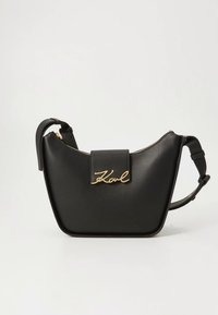 Černá kožená taška s zakřiveným tvarem, zipem na zavírání a přední klopou se zlatým podpisem. Součástí je nastavitelný popruh.