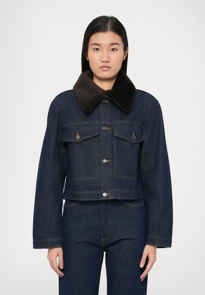 Giacca di denim blu scuro con colletto in pelliccia marrone, bottoni frontali e due tasche sul petto. Il design presenta una silhouette corta e maniche lunghe.