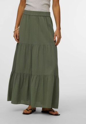 Kobieta w warstwowej oliwkowozielonej długiej spódnicy z elastycznym pasem i czarnych płaskich sandałach na jednolitym tle.