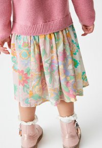 Vaaleanpunainen neulottu neule pukuun, jonka alla on värikäs kukkamekko, jossa on laskostuksia. Mekossa on vaaleanpastelleja sävyjä ja rohkeita kukkakuoseja. Vaaleanpunaiset saappaat, joissa on karvakerros.