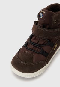 Bruna mockaskinnshöga sneakers med svarta elastiska snören, vadderad krage och vit gummisula. Guldlogotypdetalj vid vristen.