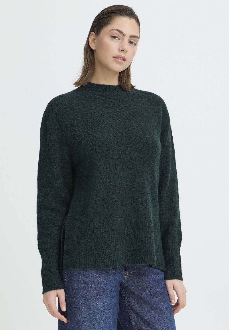 Pulover verde închis cu guler înalt, mâneci lungi, tăieturi laterale și un model tricotat texturat. Poartă cu blugi albaștri.