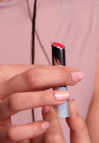 Lipstick in een metallic zilveren tube, met een heldere rode lipstick bullet. De hand houdt het schuin vast, met lichtroze nagels zichtbaar.