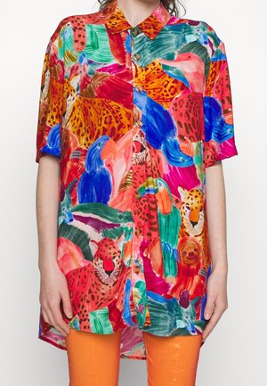 Camicetta a maniche corte con bottoni, decorata con stampe colorate di uccelli tropicali e animali, indossata con pantaloni arancioni brillanti, vista frontale dal collo alle cosce.