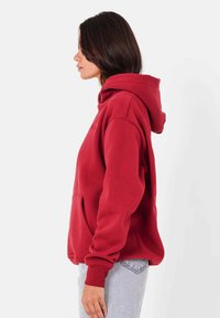 Sweat à capuche rouge en tissu doux, avec une poche avant, un cordon de serrage à la capuche et des poignets côtelés. Porté avec un jean bleu clair.