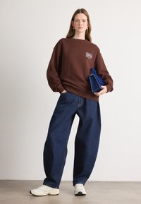 Sudadera marrón con un diseño gráfico, combinada con unos vaqueros de denim azul sueltos y zapatillas deportivas blancas, sosteniendo un pequeño bolso azul.