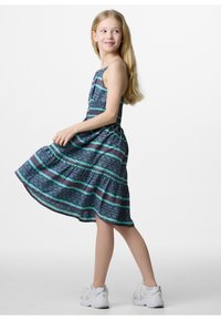 Mouwloze jurk van stoffen in blauw, groen en bordeauxrood met een patroon. A-lijn model, rok tot op de knie, gedragen met witte sportschoenen.