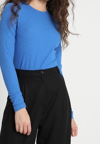 Donna che indossa una camicia blu a maniche lunghe aderente infilata in pantaloni neri a vita alta con chiusura a bottone.