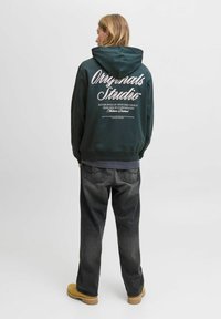 Une personne aux cheveux blonds se tient de dos, portant un sweatshirt à capuche vert foncé avec du texte blanc, un jean foncé et des bottes jaunes sur un fond uni.